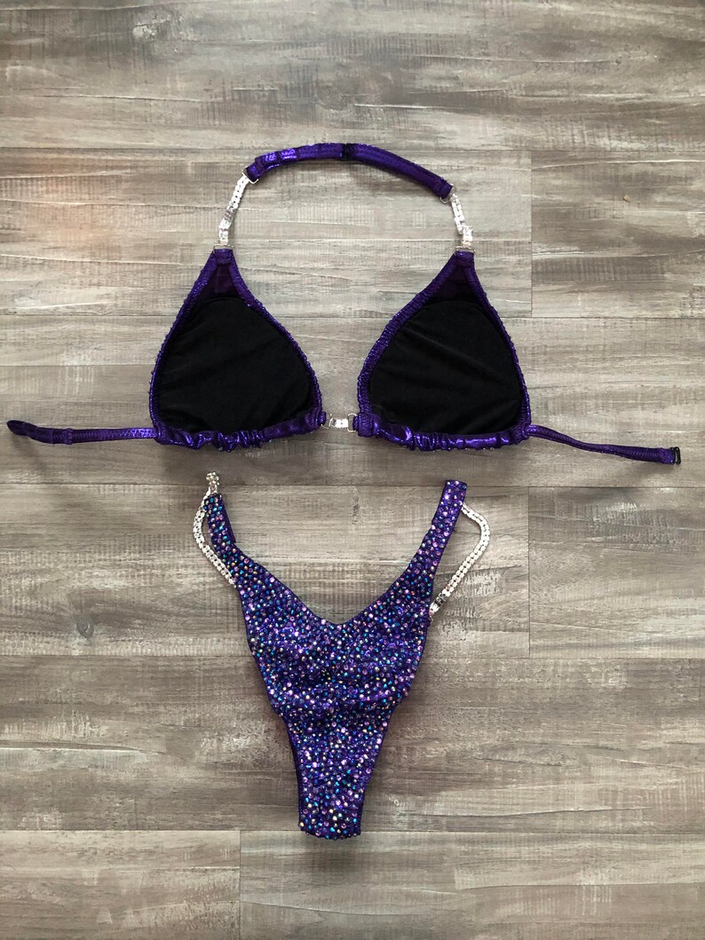 Puede incluir: Parte superior e inferior de bikini de lentejuelas moradas y plateadas. La parte superior tiene un estilo halter con un cierre en la espalda. La parte inferior tiene un corte alto y est&aacute; adornada con pedrer&iacute;a.