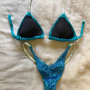 May include: A teal and black two-piece swimsuit with crystal embellishments. The top is a triangle style with adjustable straps and the bottom is a high-cut style.