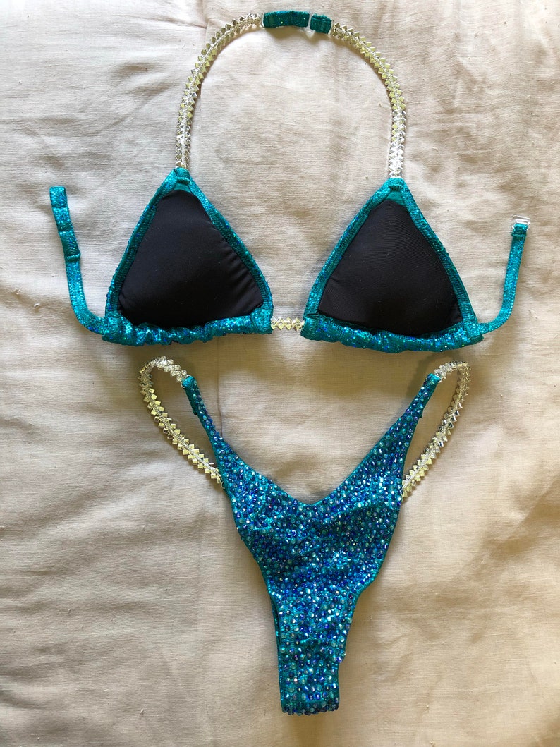 May include: A teal and black two-piece swimsuit with crystal embellishments. The top is a triangle style with adjustable straps and the bottom is a high-cut style.