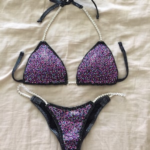 May include: A black and purple sequined bikini with crystal embellishments. The bikini top has a halter style with a tie at the back of the neck. The bikini bottom has a high-cut leg style.