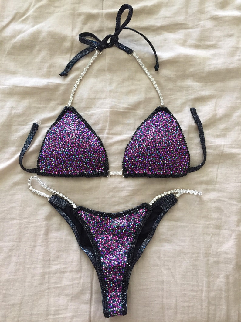 May include: A black and purple sequined bikini with crystal embellishments. The bikini top has a halter style with a tie at the back of the neck. The bikini bottom has a high-cut leg style.