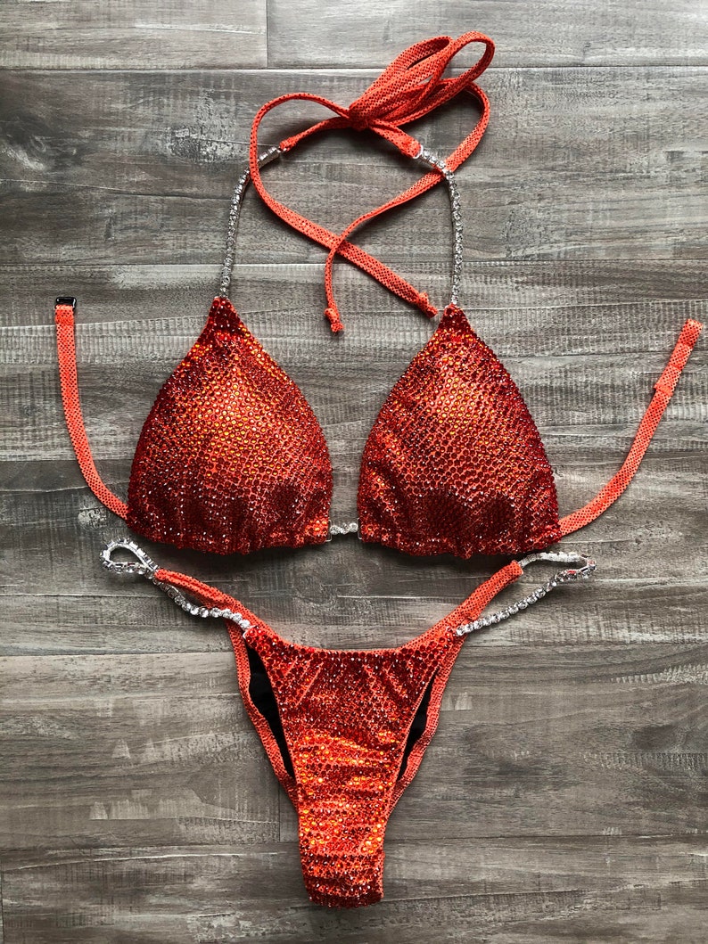 Puede incluir: Un bikini de competici&oacute;n de dos piezas con una tela brillante de color naranja intenso. La parte superior del bikini tiene forma triangular y la parte inferior tiene un estilo de corte alto.