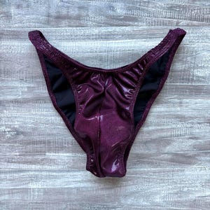 May include: A pair of purple, shiny bikini bottoms with a high-cut leg design. The bottoms have a metallic sheen and a black lining. The fabric appears to be smooth and form-fitting, suitable for swimwear or athletic wear.