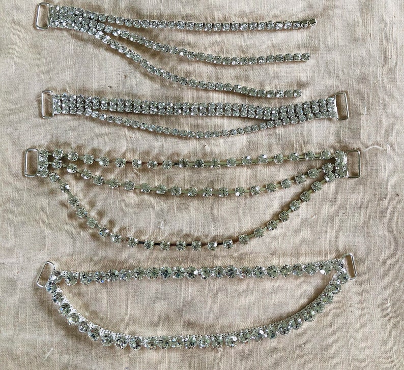 May include: Five silver rhinestone straps with clasps. The straps are arranged in a cascading pattern, with the shortest strap at the top and the longest strap at the bottom.