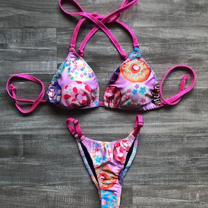 Puede incluir: Bikini rosa y morado con estampado de donut. La parte superior del bikini tiene tirantes ajustables y un estilo halter. La parte inferior del bikini tiene una pierna de corte alto.