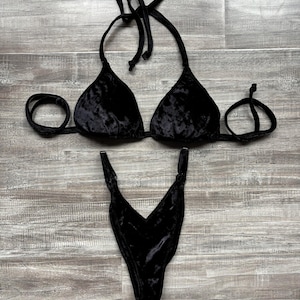 May include: A black velvet bikini set. The top is a halter style with triangle cups and tie closures. The bottom is a high-cut, V-shaped design. The fabric has a soft, textured appearance.