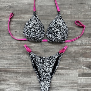 Könnte beinhalten: Ein zweiteiliger Bikini mit einem schwarz-weißen Leopardenmuster. Das Neckholder-Top und die Bikinihose sind mit leuchtend pinkfarbenen Akzenten versehen. Der Bikini wird auf einer Holzoberfläche präsentiert.