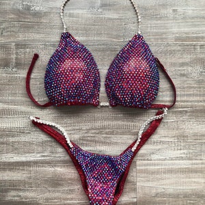 May include: A burgundy and iridescent competition bikini with crystal embellishments. The top has a halter style strap and the bottom has a high-cut leg.