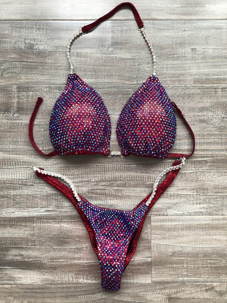 May include: A burgundy and iridescent competition bikini with crystal embellishments. The top has a halter style strap and the bottom has a high-cut leg.