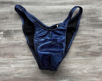 Può includere: Uno slip bikini a taglio alto realizzato in un tessuto blu scuro metallizzato e scintillante con una sottile trama glitterata. Il capo presenta una profonda scollatura a V sul davanti, sottili laccetti laterali e una fodera interna nera.