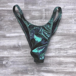 May include: A pair of high-cut bikini bottoms in a metallic emerald green fabric with a shimmering finish. The garment features a deep V-shaped front, thin side straps, and black interior lining, showcasing a sleek, form-fitting design.