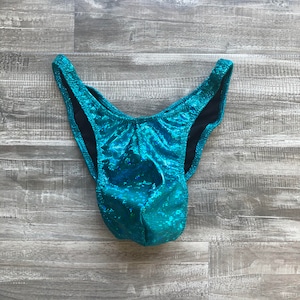 May include: A pair of high-cut bikini bottoms in a vibrant, shimmering teal fabric with a textured, reflective finish. The garment features a deep V-shaped front, high leg openings, and a solid black lining visible along the inner edges of the waistband.