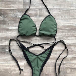 Puede incluir: Un conjunto de bikini de dos piezas en color verde oliva con ribetes negros. La parte superior tiene copas triangulares con tirantes negros al cuello, y la parte inferior es de corte alto con finas cintas negras laterales ajustables.