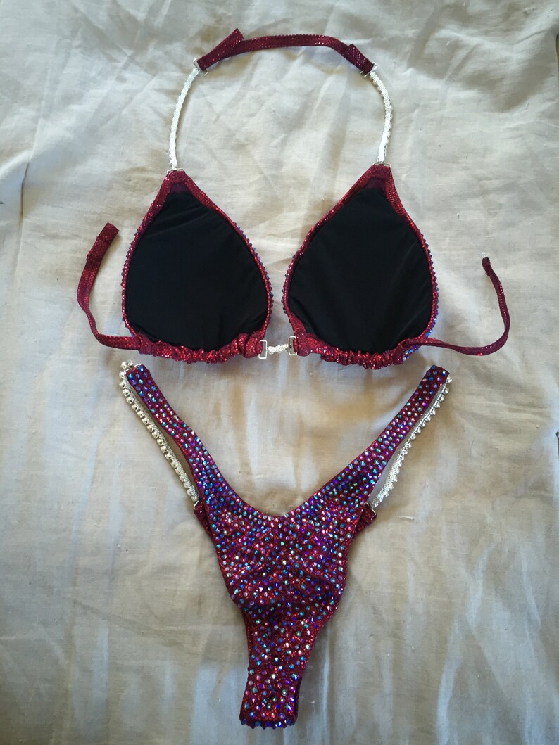 May include: A red and iridescent bikini with crystal embellishments. The top has a halter style with a white strap. The bottom is a high-cut style with a white strap on each side.