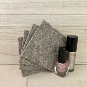 May include: A stack of five square, gray felt coasters next to two bottles of nail polish. One bottle is a light pink with glitter, and the other is a silver glitter color. Both have black caps.