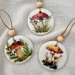 May include: Three circular ceramic ornaments featuring mushroom illustrations. Each ornament displays a different mushroom design in shades of red, brown, and orange. They are attached to a wooden bead and a brown string.