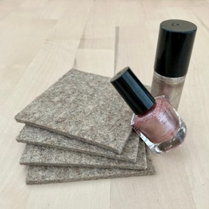May include: A stack of four square, gray felt coasters next to two bottles of nail polish. One bottle is a shimmery gold color, and the other is a pinkish-brown shade. Both bottles have black caps.