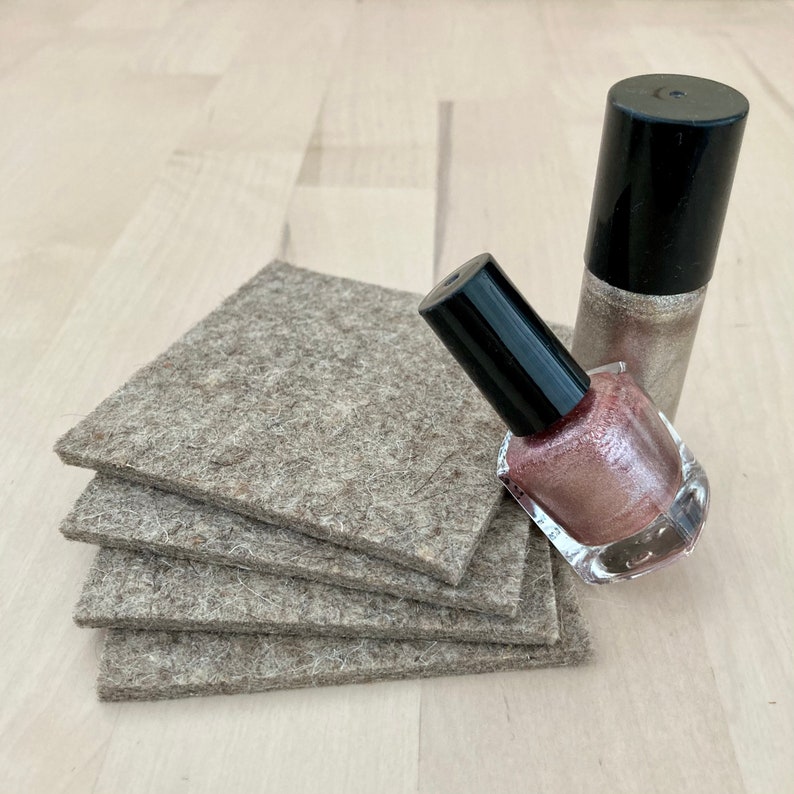 May include: A stack of four square, gray felt coasters next to two bottles of nail polish. One bottle is a shimmery gold color, and the other is a pinkish-brown shade. Both bottles have black caps.