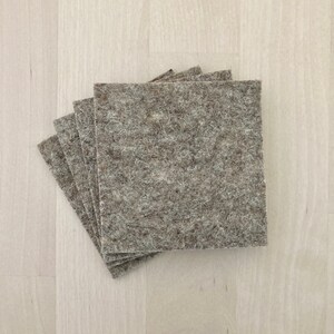 May include: A stack of five square, light gray felt pads. The pads have a textured, fibrous appearance and are arranged in a slightly fanned-out pattern, with the top pad partially obscuring the others. The felt is a neutral color.