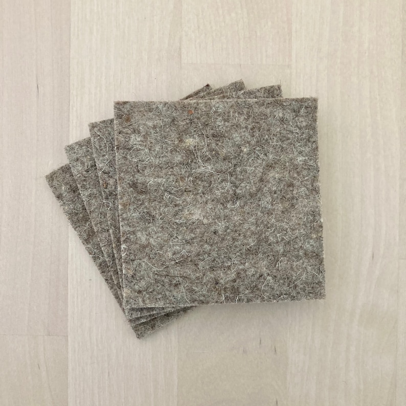 May include: A stack of five square, light gray felt pads. The pads have a textured, fibrous appearance and are arranged in a slightly fanned-out pattern, with the top pad partially obscuring the others. The felt is a neutral color.