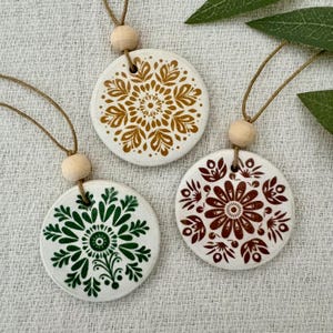 Peut inclure: Trois ornements ronds en céramique blanche avec des motifs floraux complexes en or, vert et marron. Chaque ornement est suspendu par un cordon naturel et une perle en bois. Présentés sur une surface blanche texturée.