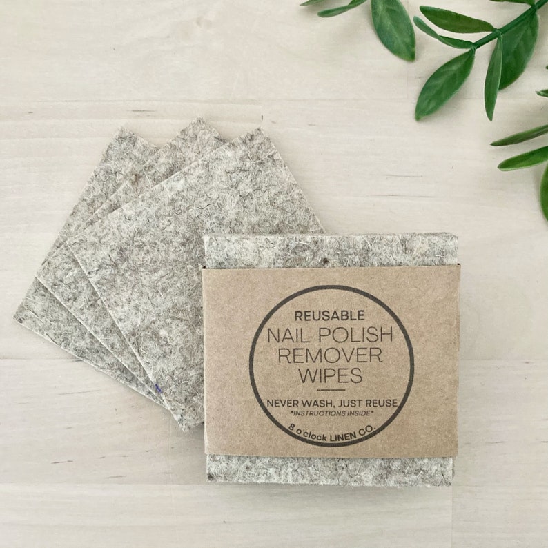 May include: A stack of reusable nail polish remover wipes made of gray fabric, with a brown paper band that reads "REUSABLE NAIL POLISH REMOVER WIPES." The wipes are square-shaped and are next to a few loose wipes.