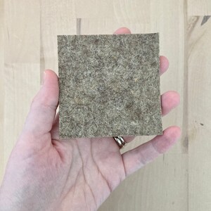 May include: A square, light brown felt pad with a textured surface. The pad is held in a hand, showcasing its size and material. The felt appears to be made of natural fibers.
