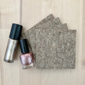 May include: Overhead shot of two nail polish bottles with black caps, one silver and one pink, next to a stack of square, gray felt pads. The items are arranged on a light wood-grain surface.