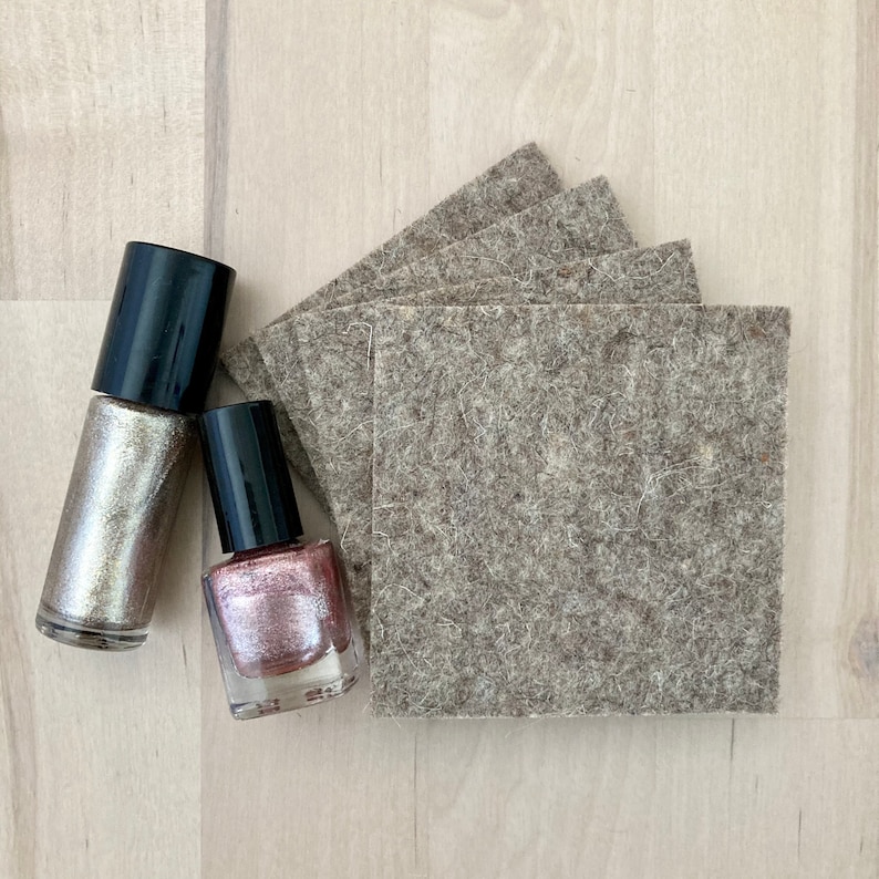 May include: Overhead shot of two nail polish bottles with black caps, one silver and one pink, next to a stack of square, gray felt pads. The items are arranged on a light wood-grain surface.