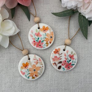 Peut inclure: Trois ornements ronds en céramique blanche avec des motifs floraux à l'aquarelle dans des tons orange, rose et vert. Chaque ornement est suspendu à une ficelle naturelle avec une perle en bois. Fleurs en arrière-plan.