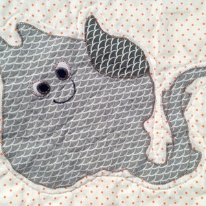 Peut inclure: Une application en tissu gris et blanc d'une raie de dessin animé avec un visage souriant et de grands yeux. La raie est sur un fond blanc avec des pois orange.