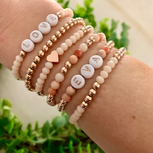 May include: A set of six bracelets with white beads and gold accents. The bracelets feature the letters "LOVE" and "EVA" in gold lettering on white beads. One bracelet has a small gold heart charm.