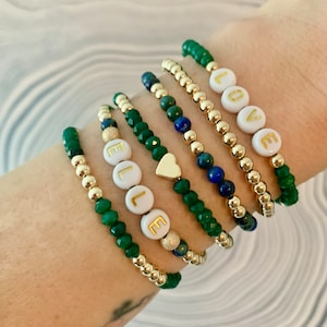 May include: A set of six bracelets with gold beads and green and blue gemstone beads. The bracelets feature white beads with gold lettering spelling out "LOVE" and "ELLE". One bracelet has a gold heart charm.