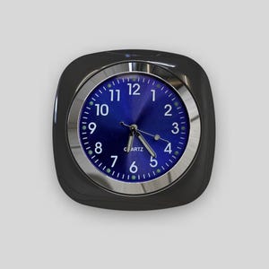 May include: A black square clock with a silver rim and a blue face. The clock has white numbers and hands, and the word "QUARTZ" is printed in white on the face.
