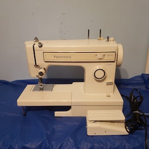 Puede incluir: Una máquina de coser Kenmore blanca con un pedal y un cable de alimentación. La máquina tiene un cuerpo de plástico blanco y una placa de aguja de metal plateado. La máquina tiene una variedad de ajustes de puntada y un enhebrador de aguja incorporado.