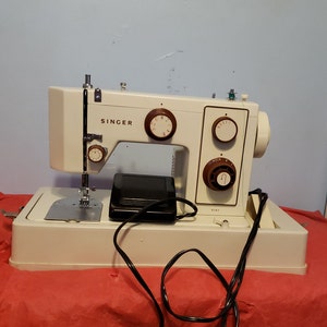 May include: A white Singer sewing machine with a black foot pedal and a power cord. The machine has a brown plastic casing and several dials. The machine is sitting on a red surface.