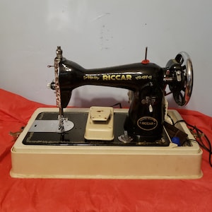 May include: A black vintage Riccar sewing machine with a white base and a foot pedal. The machine has a gold-colored nameplate with the word "Riccar" on it.