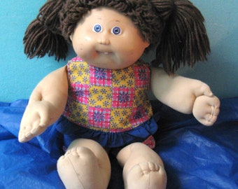 Puede incluir: Una muñeca vintage con rostro de vinilo, ojos azules y cabello castaño de hilo en coletas. La muñeca viste un top estampado con cuadros rosas, amarillos y azules, y una falda azul. Está sentada sobre una superficie azul.