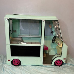 Puede incluir: Un camión de helados de juguete verde menta con una ventana negra y un interior rosa y blanco. El camión tiene un letrero negro en el lateral que dice "OGH!"
