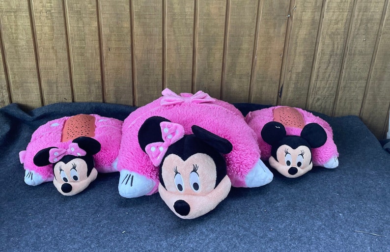 Pu&ograve; includere: Tre cuscini rosa in peluche del personaggio di Minnie Mouse. Ogni cuscino presenta il volto di Minnie Mouse, orecchie nere e un fiocco. I cuscini sono su una superficie grigia con uno sfondo in legno.