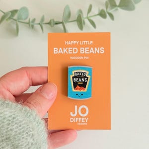 May include: A wooden pin shaped like a blue can of baked beans with a smiling face, mounted on an orange display card. The card features the text HAPPY LITTLE BAKED BEANS WOODEN PIN and JO DIFFEY DESIGNS. The pin graphic shows a black label.