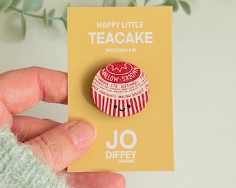 May include: A circular wooden pin featuring a red and white striped Tunnock's Teacake design with a small smiling face. The pin is attached to a yellow display card printed with the text HAPPY LITTLE TEACAKE WOODEN PIN and JO DIFFEY DESIGNS.