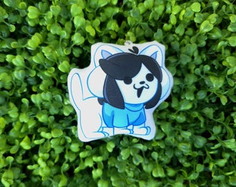 May include: A cartoon-style keychain featuring a character with black hair, a blue outfit, and a white outline. The keychain is set against a backdrop of green foliage. The character has a smiling expression.