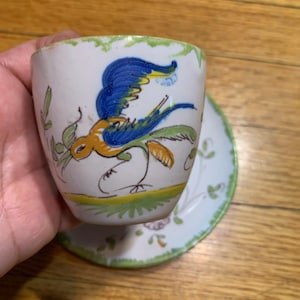 May include: A white ceramic cup and saucer set featuring a hand-painted design of a blue and yellow bird in flight. The rim of both pieces is decorated with a green scalloped pattern, and the saucer includes additional floral motifs in soft colors.