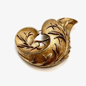 May include: A gold-toned brooch in the shape of a stylized leaf with intricate details. The brooch is made of metal and has a textured surface.