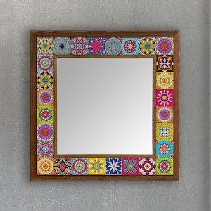 Może przedstawiać: Kwadratowe lustro w drewnianej ramie z kolorowymi, wzorzystymi kafelkami na obrzeżach. Kafelki przedstawiają skomplikowane wzory kwiatowe i geometryczne w odcieniach różu, niebieskiego, żółtego i fioletowego. Dekoracyjne lustro ścienne.
