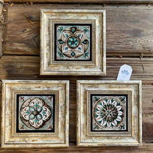 Può includere: Tre piastrelle quadrate incorniciate con disegni ornati turchese, marrone e bianco. Le cornici hanno una finitura bianco sporco anticata. Le piastrelle sono su uno sfondo di legno rustico.