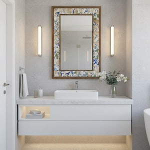 以下が含まれることがあります： 装飾的なモザイクタイルのフレームが付いた大きな鏡のあるモダンなバスルーム。白い洗面化粧台には長方形のシンクがあり、床には青いラグがあります。2つの縦型ライトが鏡の両側にあり、花瓶の花がエレガントさを添えています。