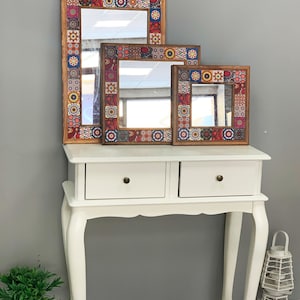 May include: Three decorative mirrors with wooden frames and colorful mosaic tile borders. The mirrors are rectangular and square, varying in size, and are displayed on a white console table with two drawers. The mosaic tiles feature a mix of red, blue, yellow, and brown patterns.