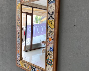 May include: A rectangular wall mirror featuring a wooden frame bordered by a mosaic of colorful patterned tiles. The tiles display various geometric and floral designs in shades of blue, yellow, orange, and white. The mirror reflects a storefront.